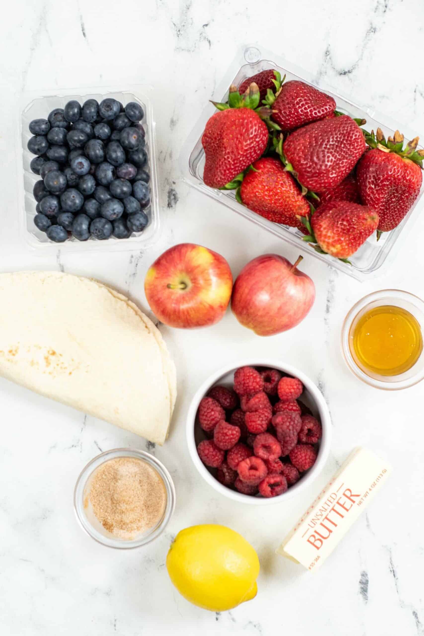 A selection of fruits and ingredients on a white surface: blueberries, strawberries, apples, raspberries, honey, a lemon, brown sugar, butter.