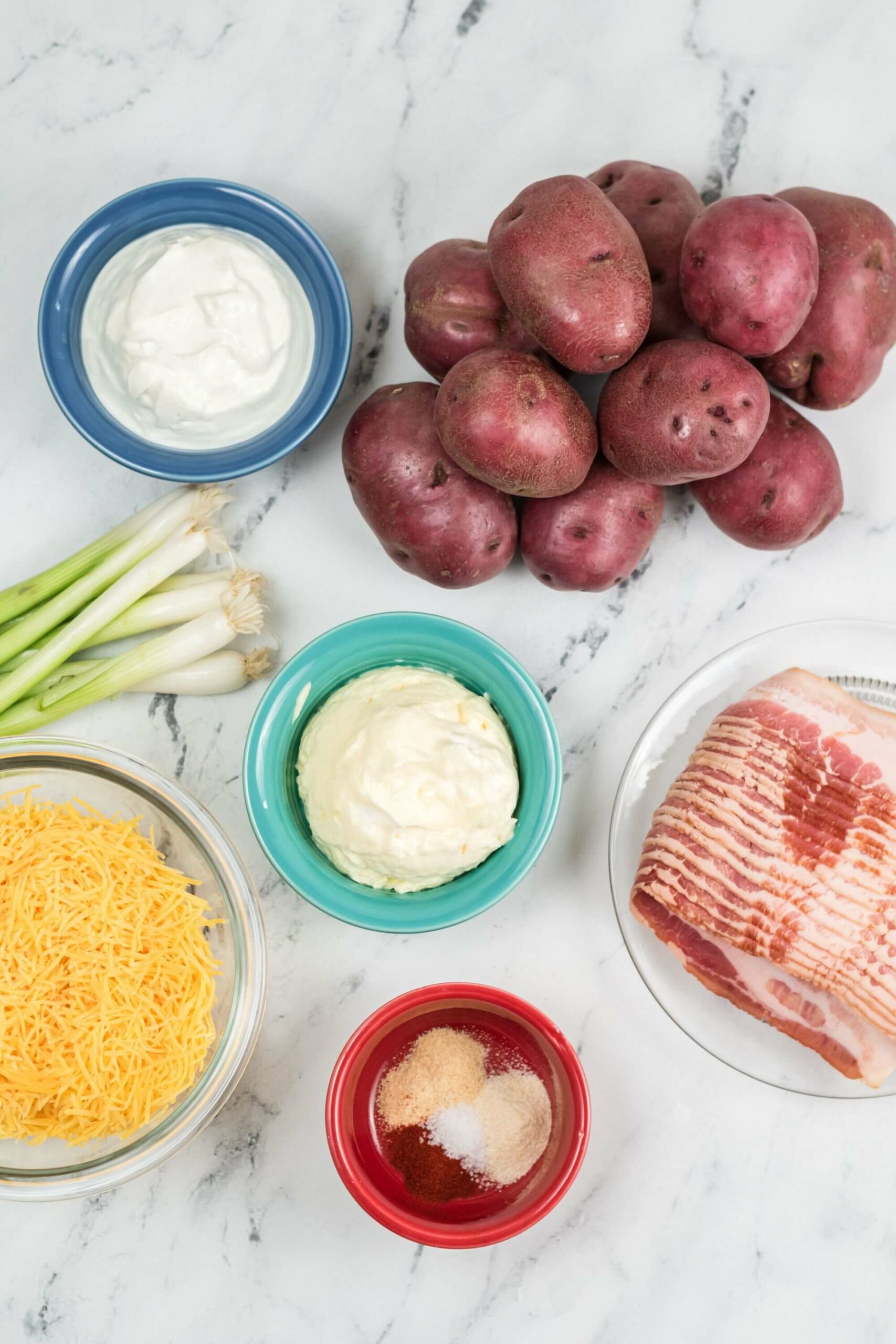 Ingredients for a Loaded Baked Potato Salad recipe on a marble countertop, including bowls of sour cream, mayonnaise, cheese, spices, green onions, potatoes, and bacon.