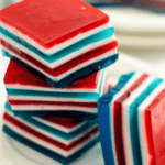Three vibrant, layered Jello squares in red, white, and blue are stacked on a plate, with more similar squares in the background.