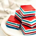 A plate of layered Jello desserts with red, white, and blue stripes is shown, placed on a white surface with a blurred background.