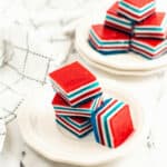 Stacked red, white, and blue layered Jello squares rest on two white plates, with a white cloth featuring a black grid pattern next to them.