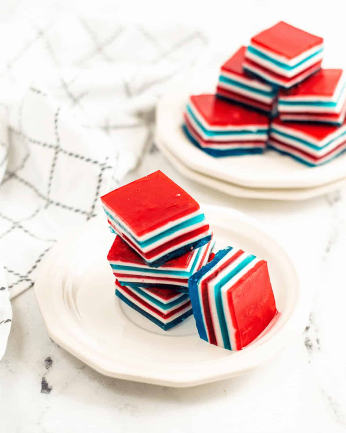 Stacked red, white, and blue layered Jello squares rest on two white plates, with a white cloth featuring a black grid pattern next to them.