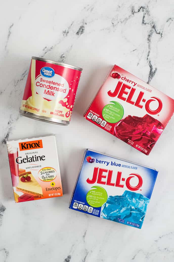 A can of sweetened condensed milk and three boxes of gelatin mix (gelatine, cherry Jell-O, berry blue Jell-O) are arranged on a marble surface, ready to create a delightful layered jello dessert.