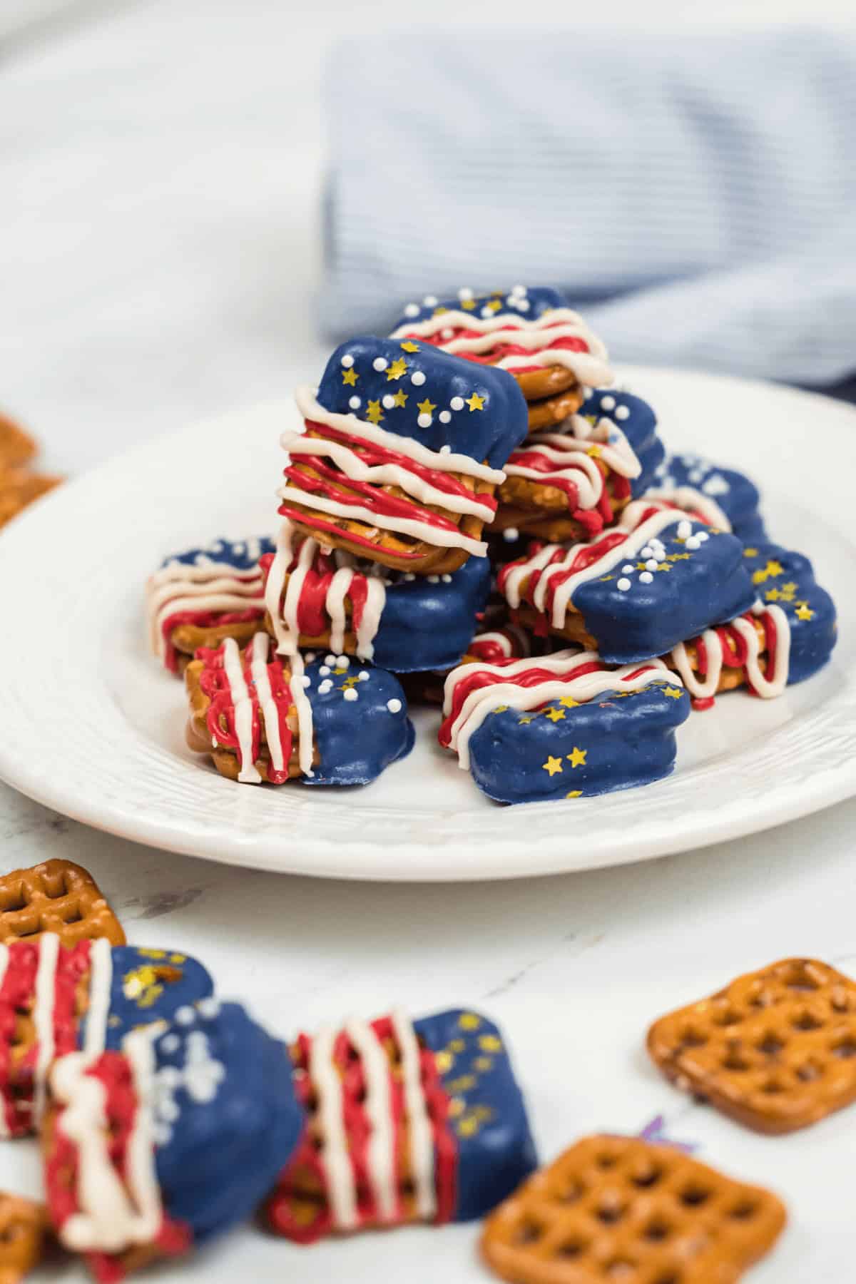 A plate covered with chocolate, dipped in blue icing and adorned with white and red drizzles, decorated with white and yellow sprinkles. Additional pretzels are scattered around the plate, showcasing a festive treat.