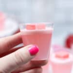 A hand with pink nail polish holds a clear cup filled with pink liquid and two small pink cubes on top, resembling delightful Strawberry Jello Shots.