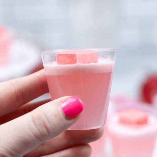 A hand with pink nail polish holds a clear cup filled with pink liquid and two small pink cubes on top, resembling delightful Strawberry Jello Shots.