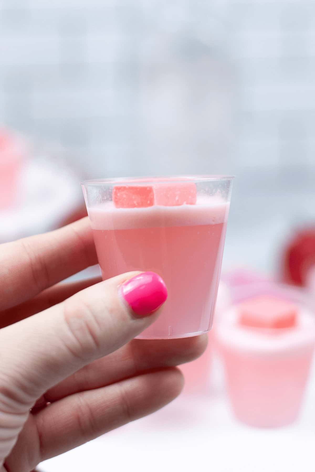 A hand with pink nail polish holds a clear cup filled with pink liquid and two small pink cubes on top, resembling delightful Strawberry Jello Shots.