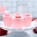 Clear cups are displayed on a white scalloped cake stand. A pink candy piece tops each shot, with more candies in the background.