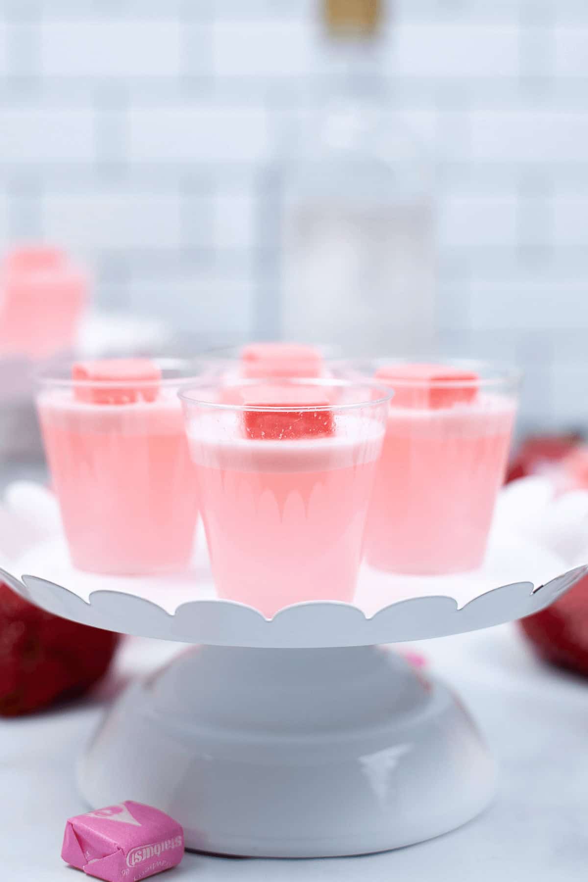 Clear cups are displayed on a white scalloped cake stand. A pink candy piece tops each shot, with more candies in the background.