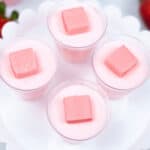 Four small clear cups filled with pink mousse, each topped with a pink square of candy, arranged on a white scalloped tray. Strawberries are partially visible in the background, giving a nod to delightful Strawberry Jello Shots.