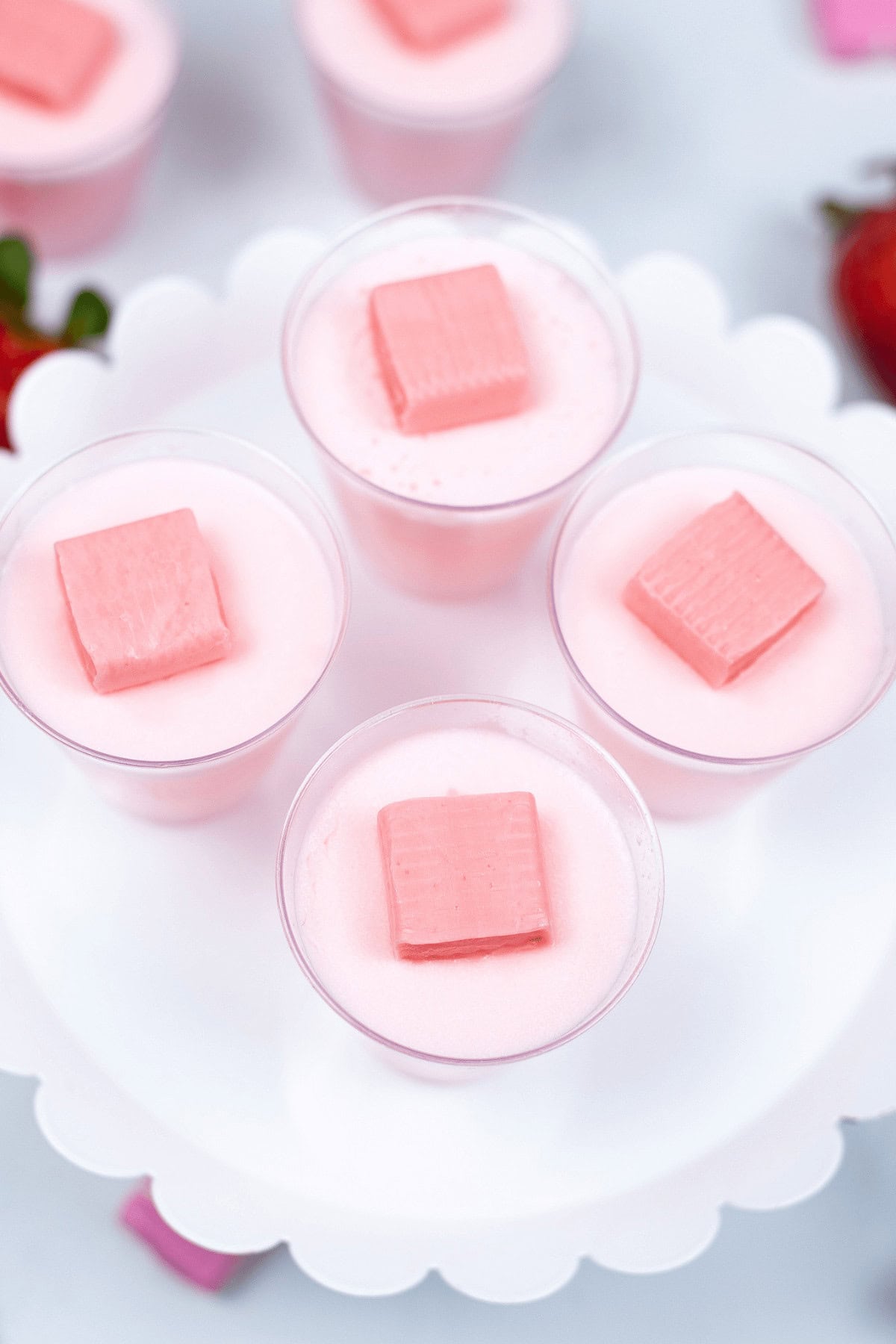 Four small clear cups filled with pink mousse, each topped with a pink square of candy, arranged on a white scalloped tray. Strawberries are partially visible in the background, giving a nod to delightful Strawberry Jello Shots.