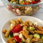 A bowl of Rotini Pasta Salad with cherry tomatoes, cucumbers, red onions, mozzarella cubes, and mixed seasonings. A glass bowl containing more salad is in the background.