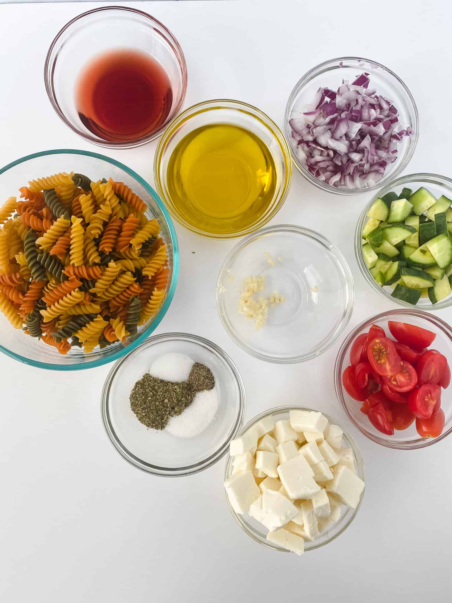 Bowls containing ingredients for Rotini Pasta Salad: tricolor rotini pasta, red wine vinegar, olive oil, chopped red onions, chopped cucumbers, minced garlic, halved cherry tomatoes, feta cheese cubes, and a mix of seasoning.