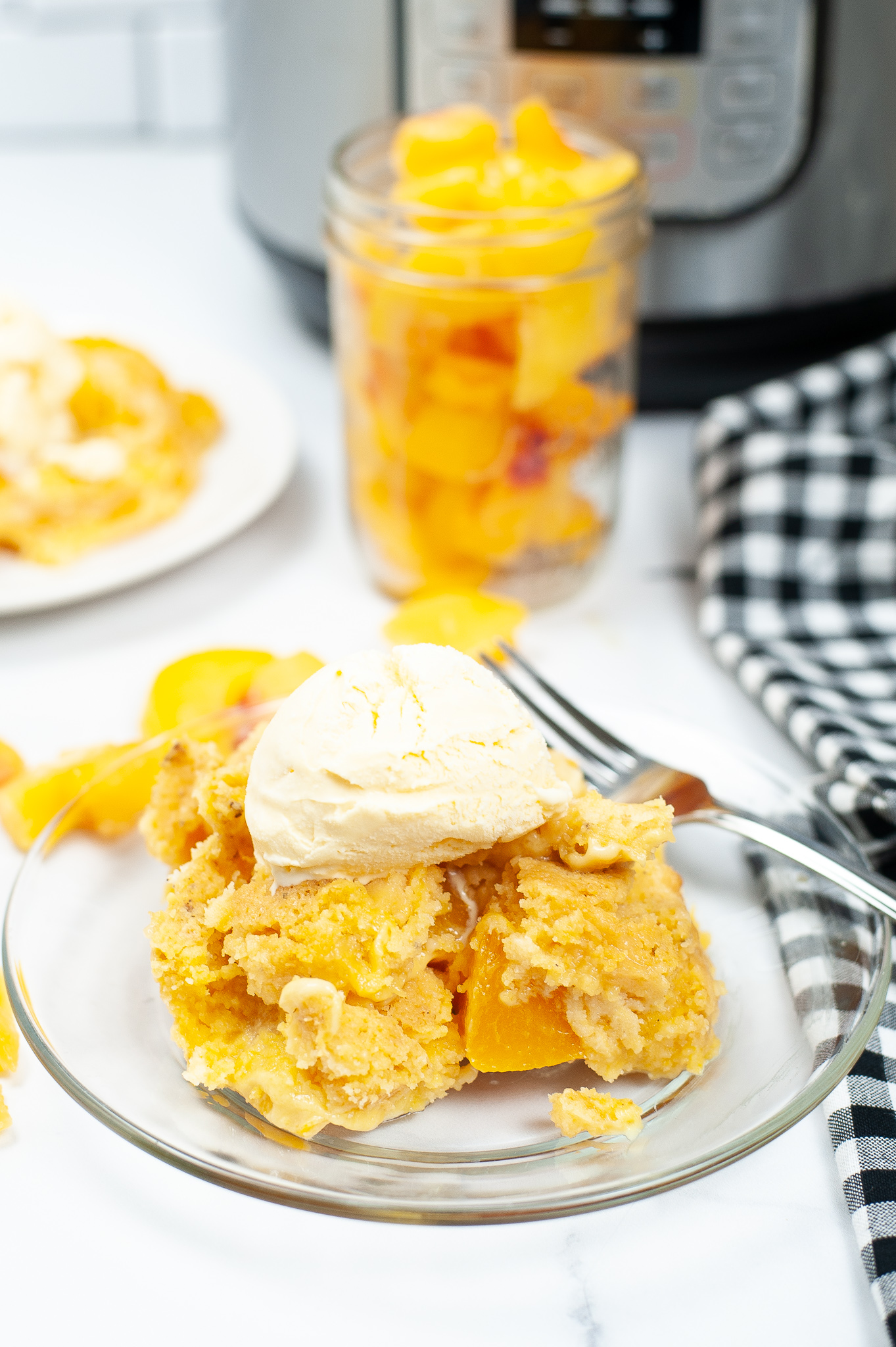 Peach cobbler on a clear plate with ice cream and peaches in a jar in the background.