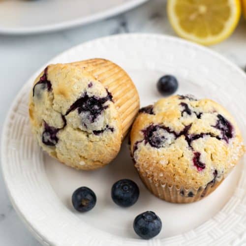Two blueberry muffins on a white plate with a scattering of blueberries on the top.