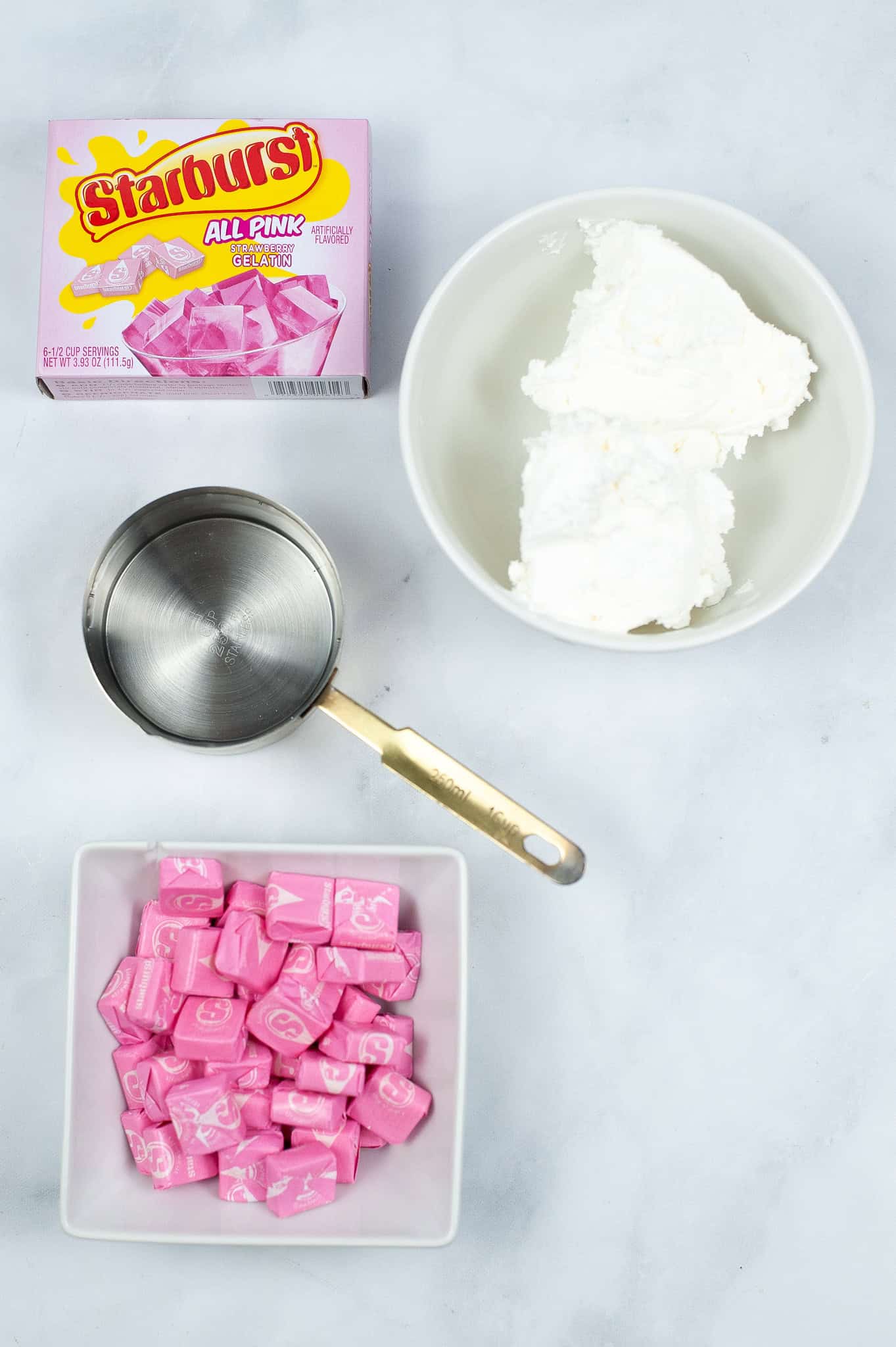 Top view of a food preparation scene featuring a box of Starburst candy, a bowl of white cream, a measuring cup, and a bowl of pink Starburst candies, perfect for making Strawberry Jello Shots.