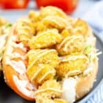 A shrimp po boy sandwich with fried shrimp on lettuce and tomato slices, drizzled with sauce, rests on a gray plate. Juicy tomatoes linger in the background.
