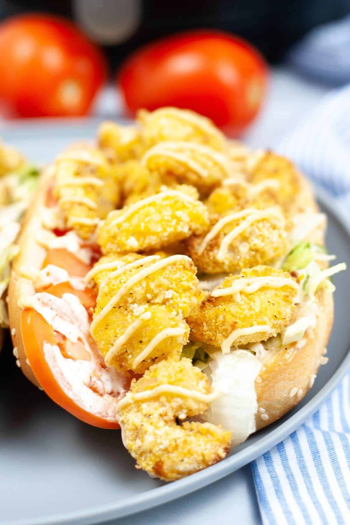 A shrimp po boy sandwich with fried shrimp on lettuce and tomato slices, drizzled with sauce, rests on a gray plate. Juicy tomatoes linger in the background.