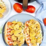 Two sandwiches with fried chicken, lettuce, and tomato on a gray plate. Reminiscent of a shrimp po boy sandwich, they're drizzled with sauce. An air fryer, two whole tomatoes, and a blue-striped cloth sit in the background.