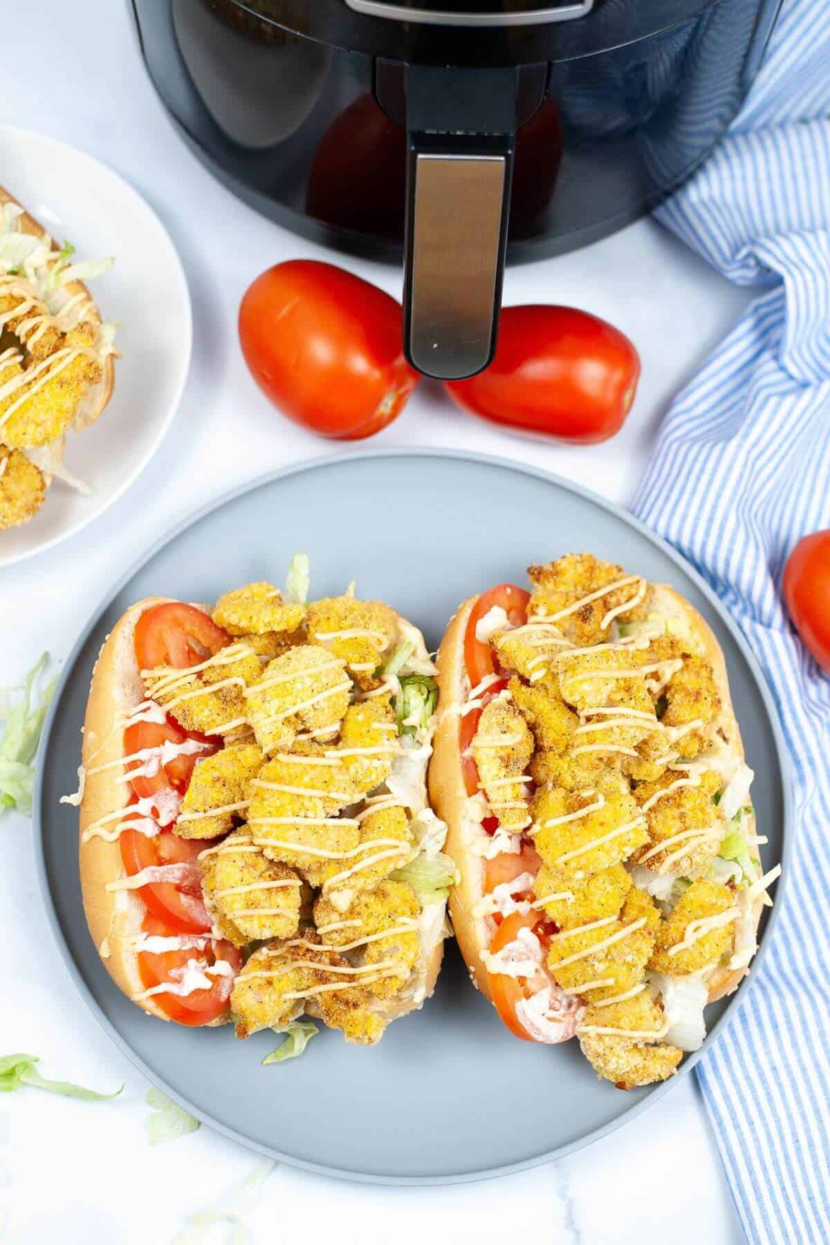 Two sandwiches with fried chicken, lettuce, and tomato on a gray plate. Reminiscent of a shrimp po boy sandwich, they're drizzled with sauce. An air fryer, two whole tomatoes, and a blue-striped cloth sit in the background.