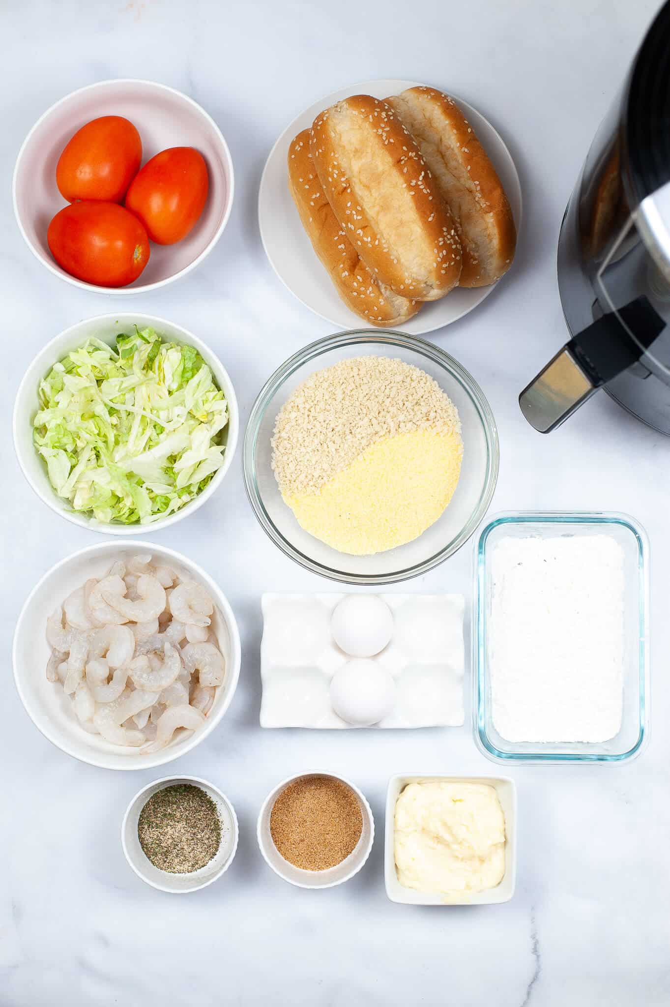 Ingredients are arranged on the countertop: tomatoes, buns, shredded lettuce, succulent shrimp, eggs, breadcrumbs, flour, seasonings, and mayonnaise alongside an air fryer.