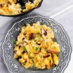 A serving of cheesy potato casserole with bacon pieces and herbs on a decorative gray plate, with a hearty Cast Iron Skillet Breakfast casserole in the background.