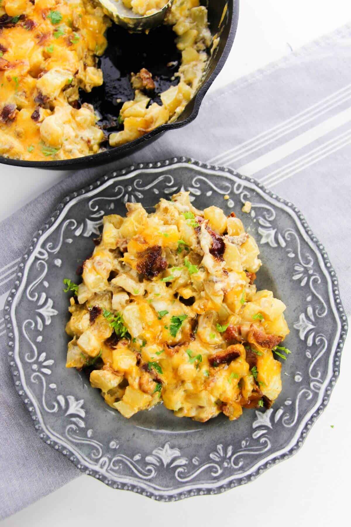 A serving of cheesy potato casserole with bacon pieces and herbs on a decorative gray plate, with a hearty Cast Iron Skillet Breakfast casserole in the background.