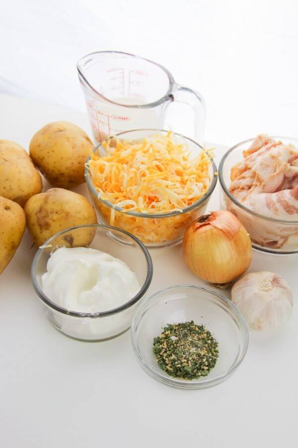 Assorted ingredients on a white surface for a Cast Iron Skillet Breakfast: potatoes, shredded cheese, bacon, an onion, garlic, a bowl of sour cream, mixed herbs, and a measuring cup with liquid.
