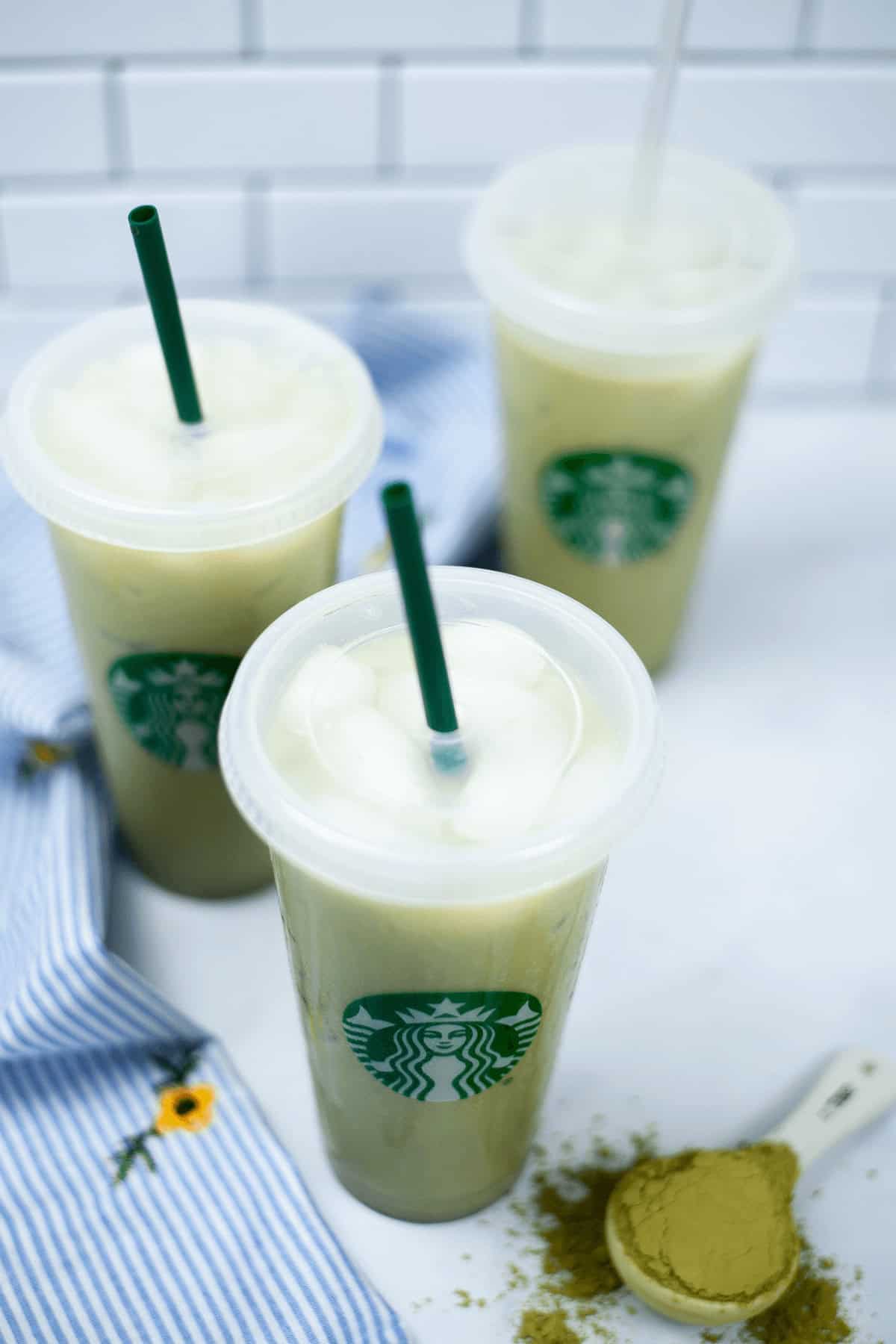 Three iced matcha lattes, nestled in iconic Starbucks cups, sit on a white surface adorned with a blue striped cloth. Nearby, a scoop of vibrant matcha powder has artistically spilled, inviting you to savor the refreshing charm of an iced matcha latte.