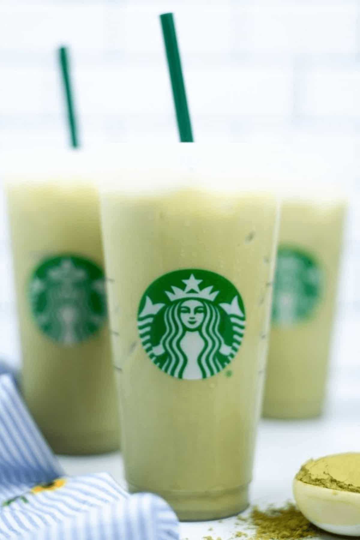 Three iced matcha lattes in clear Starbucks cups with green straws are shown. A spoon with rich, vibrant matcha powder is visible in the foreground, highlighting the refreshing appeal of the iced matcha latte experience.