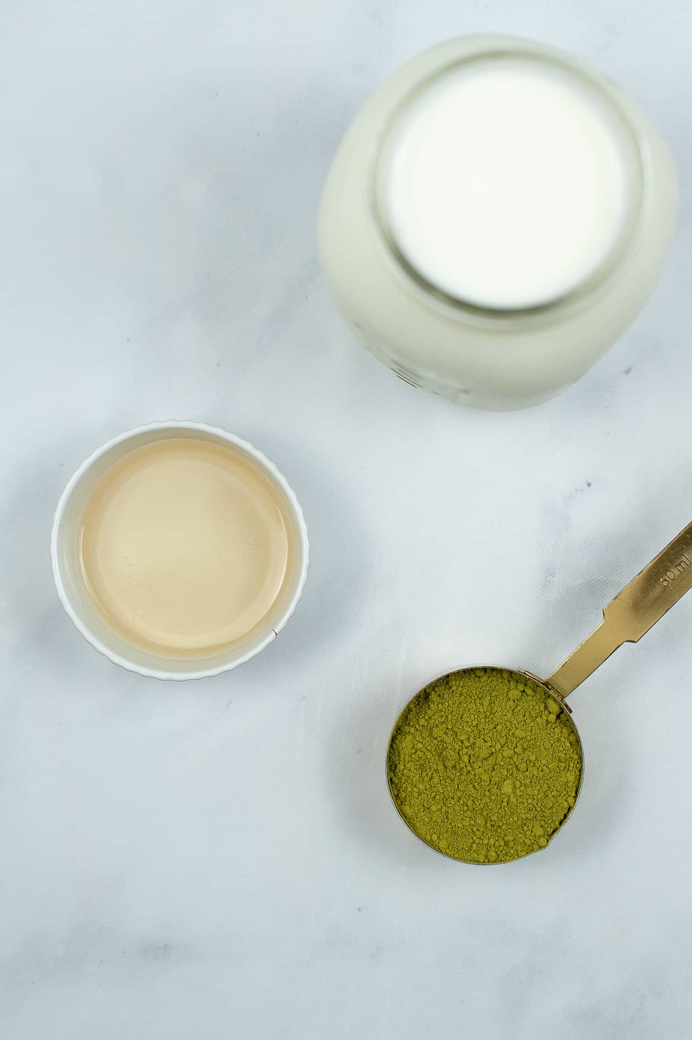 A cup of matcha powder, a jar with milk, and a small bowl with liquid rest on a white surface, ready to craft the perfect iced matcha latte.