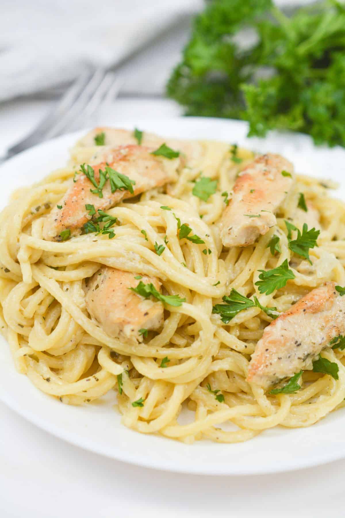 A plate of creamy chicken pesto pasta topped with tender pieces of cooked chicken and garnished with chopped parsley.