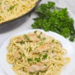 A plate of chicken pesto pasta with creamy sauce and grilled chicken pieces, garnished with chopped parsley, sits next to a skillet and a bunch of fresh parsley.