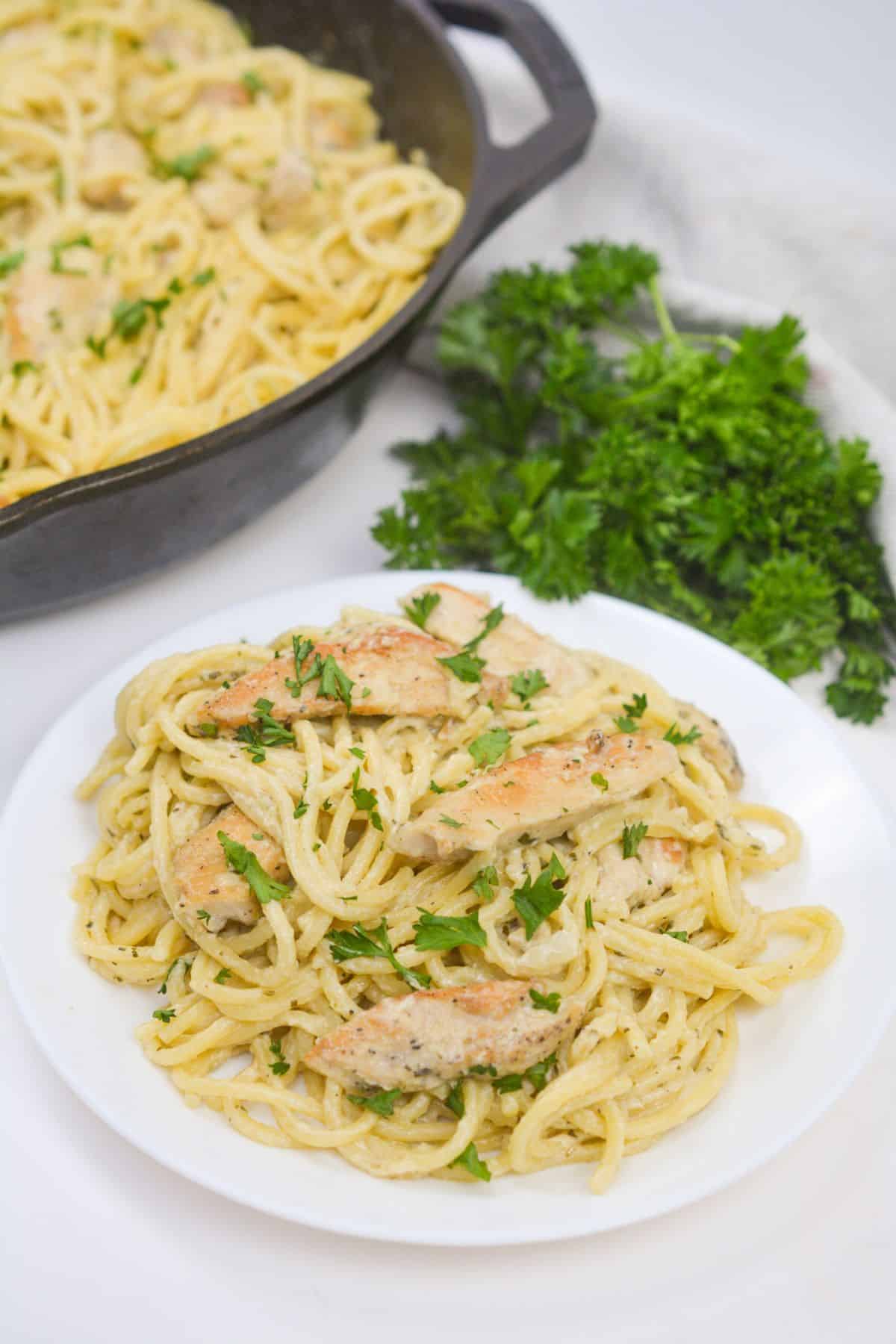 A plate of chicken pesto pasta with creamy sauce and grilled chicken pieces, garnished with chopped parsley, sits next to a skillet and a bunch of fresh parsley.