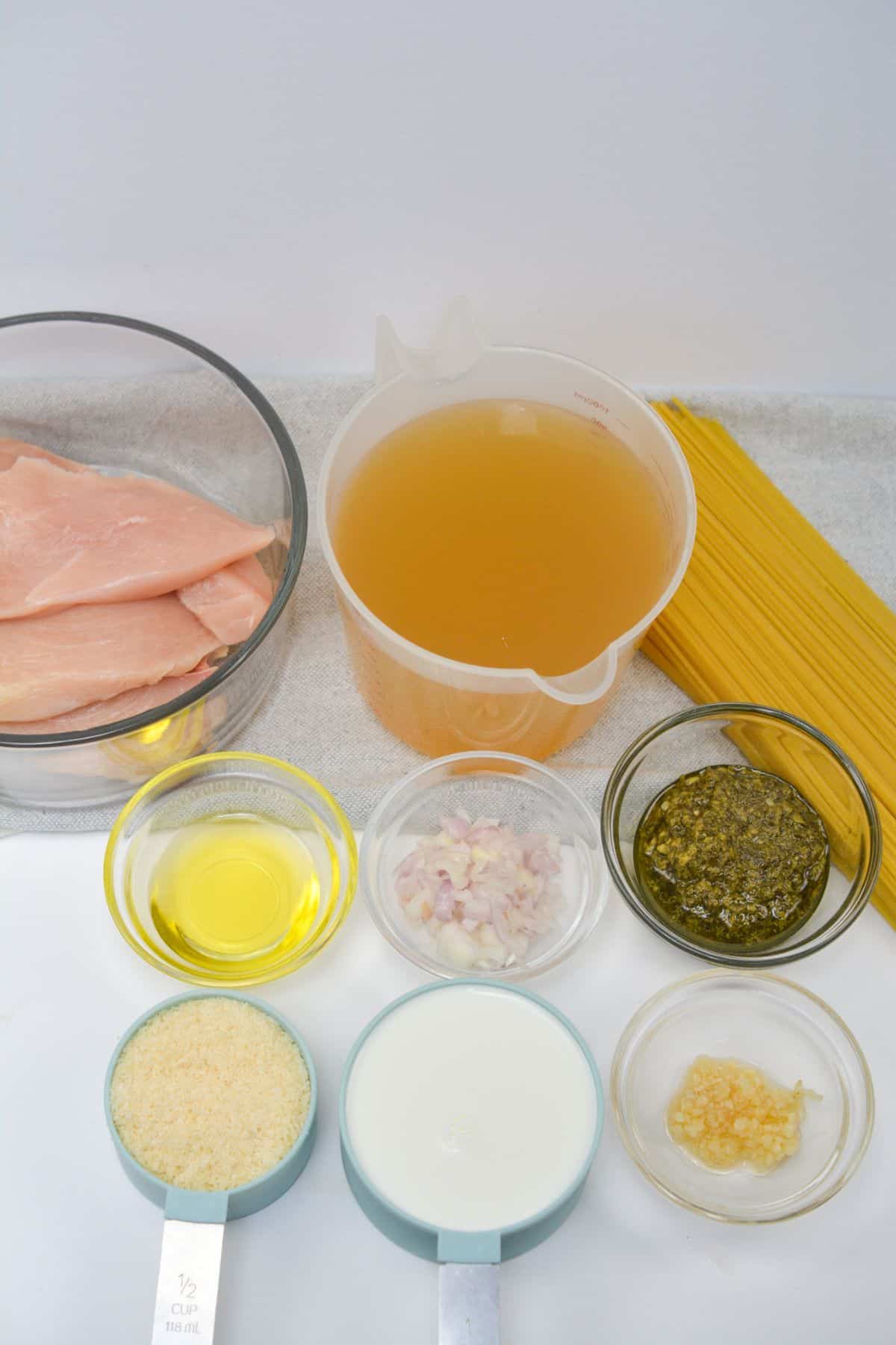 Ingredients raw chicken breasts, a jug of chicken broth, uncooked spaghetti, olive oil, minced shallot, pesto, minced garlic, grated Parmesan, and milk arranged on a countertop.
