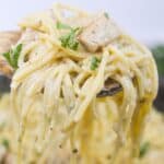 A close-up of a fork lifting a serving of creamy chicken pesto pasta with pieces of chicken and garnished with fresh parsley.