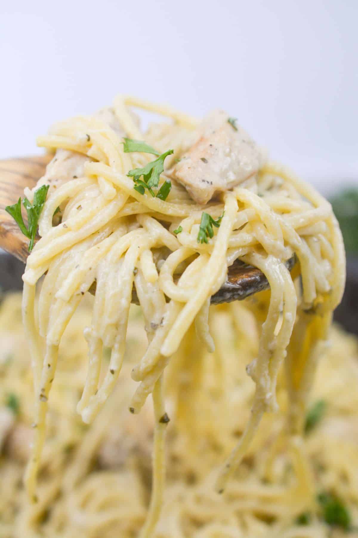 A close-up of a fork lifting a serving of creamy chicken pesto pasta with pieces of chicken and garnished with fresh parsley.