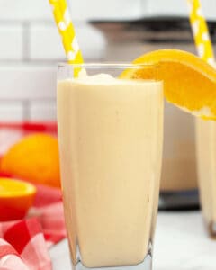 A glass of light of an Orange Creamsicle Milkshake, with an orange slice and a yellow polka dot straw, set against a blurred background with another glass and some whole oranges.