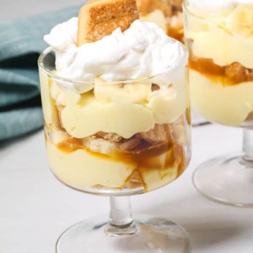 An easy banana pudding delight: a glass dessert cup layered with creamy pudding, banana slices, caramel sauce, and fluffy whipped cream, all crowned with a cookie. Two similarly tempting cups linger in the background.