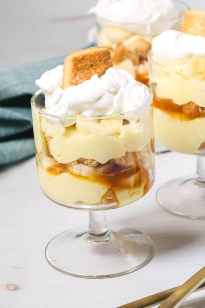 An easy banana pudding delight: a glass dessert cup layered with creamy pudding, banana slices, caramel sauce, and fluffy whipped cream, all crowned with a cookie. Two similarly tempting cups linger in the background.