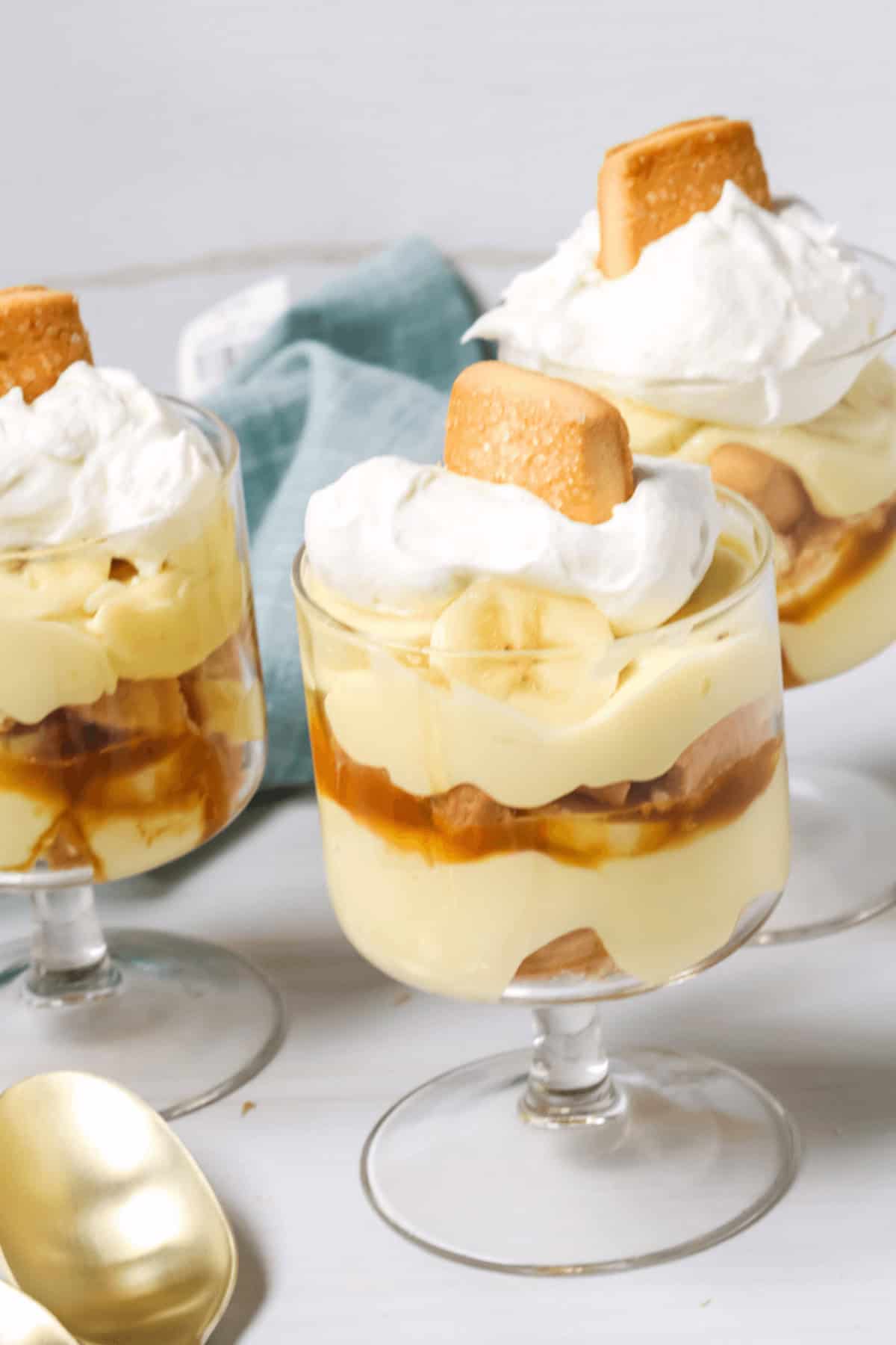 Three glass dishes of layered, dessert topped with whipped cream and a cookie, next to a blue napkin and a gold spoon.