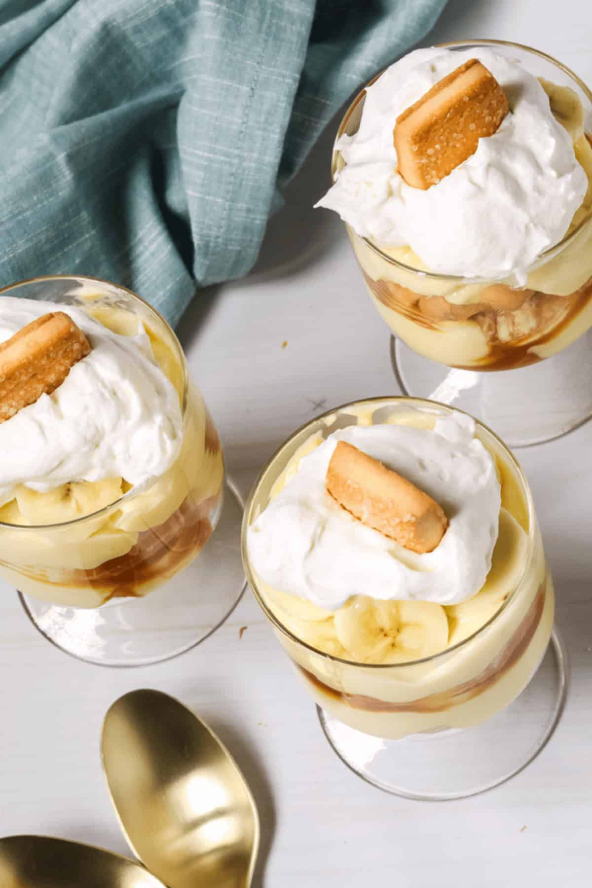 Three glasses of easy banana pudding topped with whipped cream and a cookie rest on a white surface. A blue cloth drapes beside them, along with two gold spoons, ready to scoop up the deliciousness.