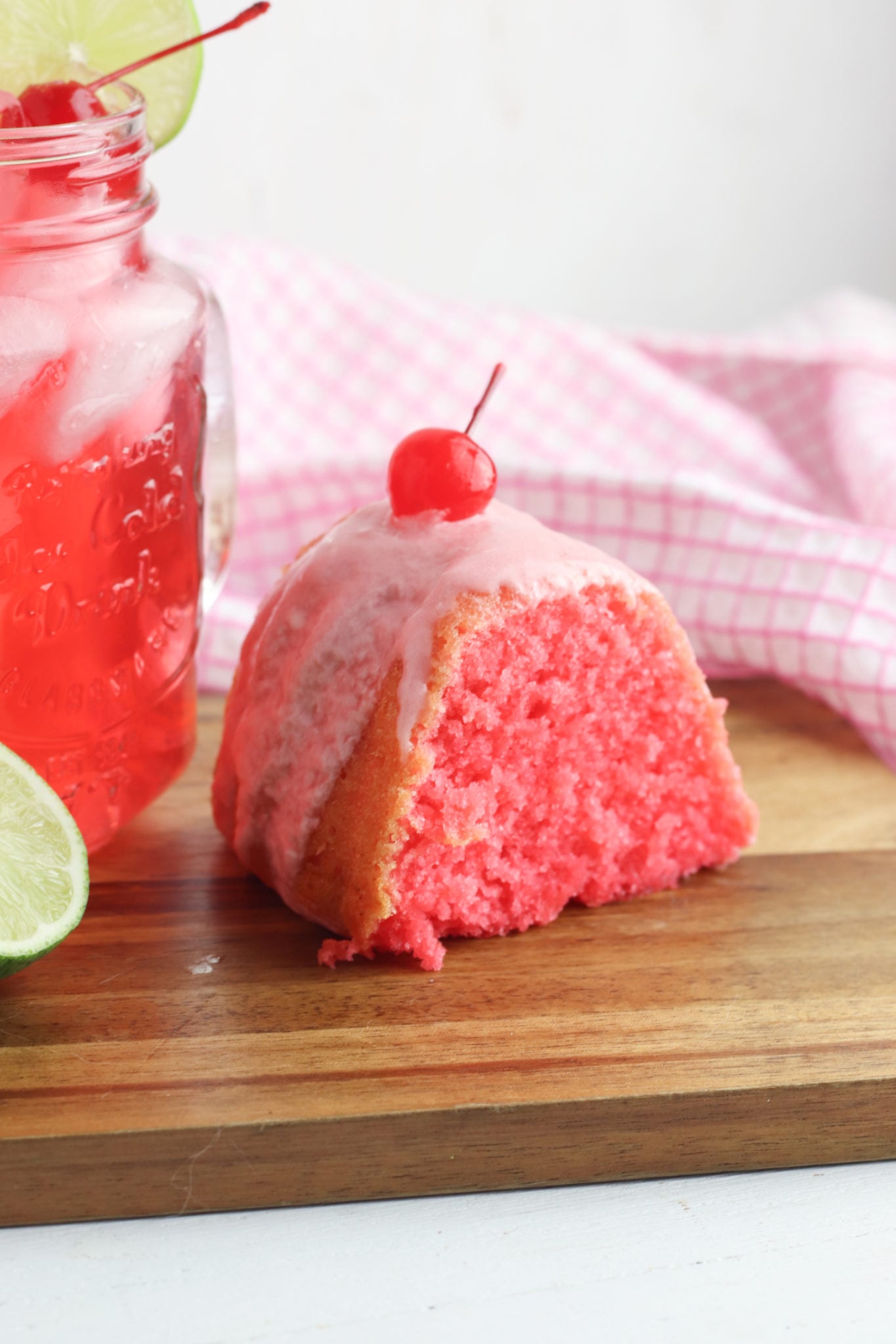 Piece of cherry limeade with a cherry on top and a glass of cherry limeade beside t.