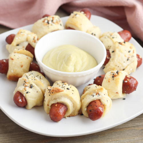 A ring of crescent pigs in a blanket around dipping sauce on a white plate.