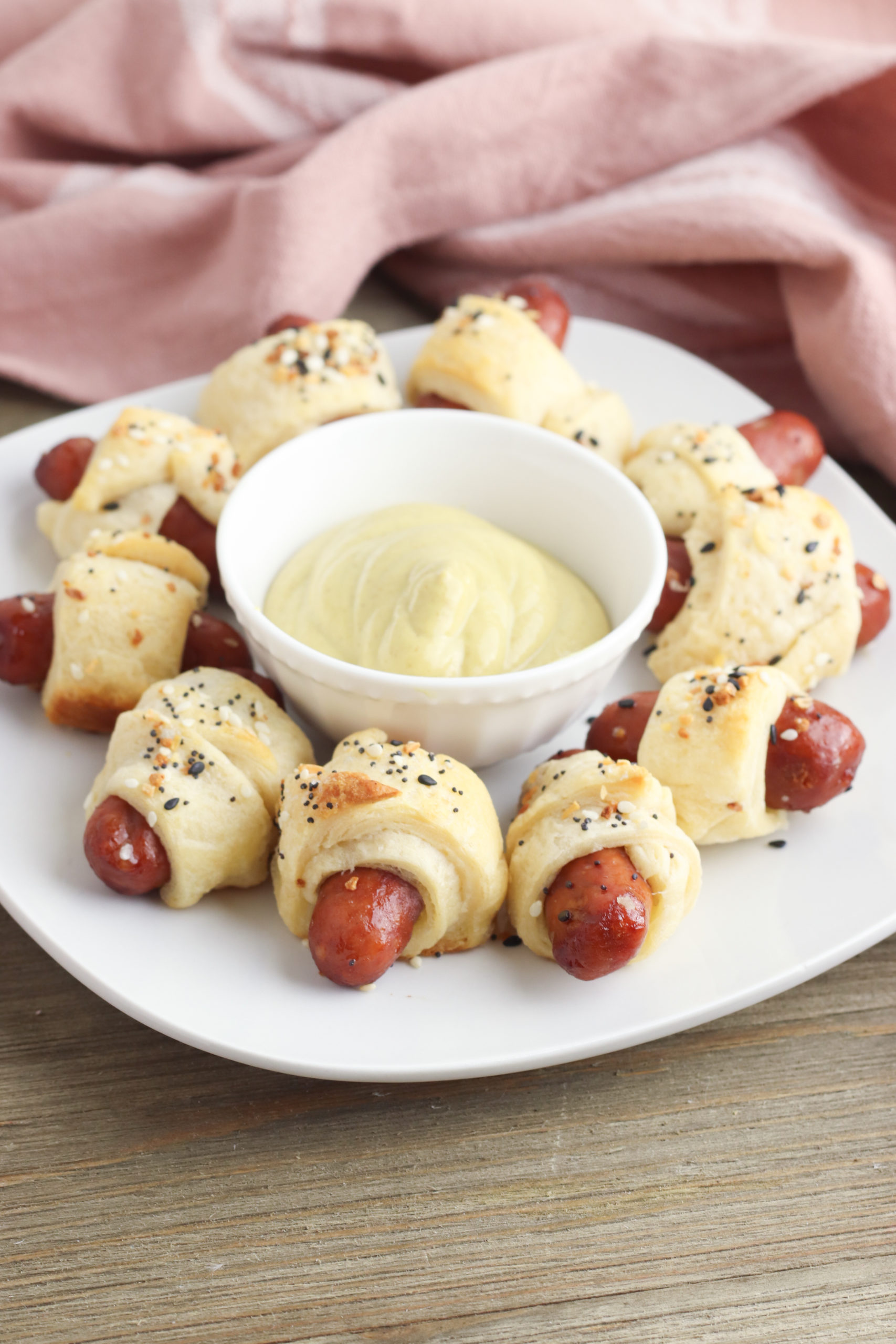 A ring of crescent pigs in a blanket around dipping sauce on a white plate.