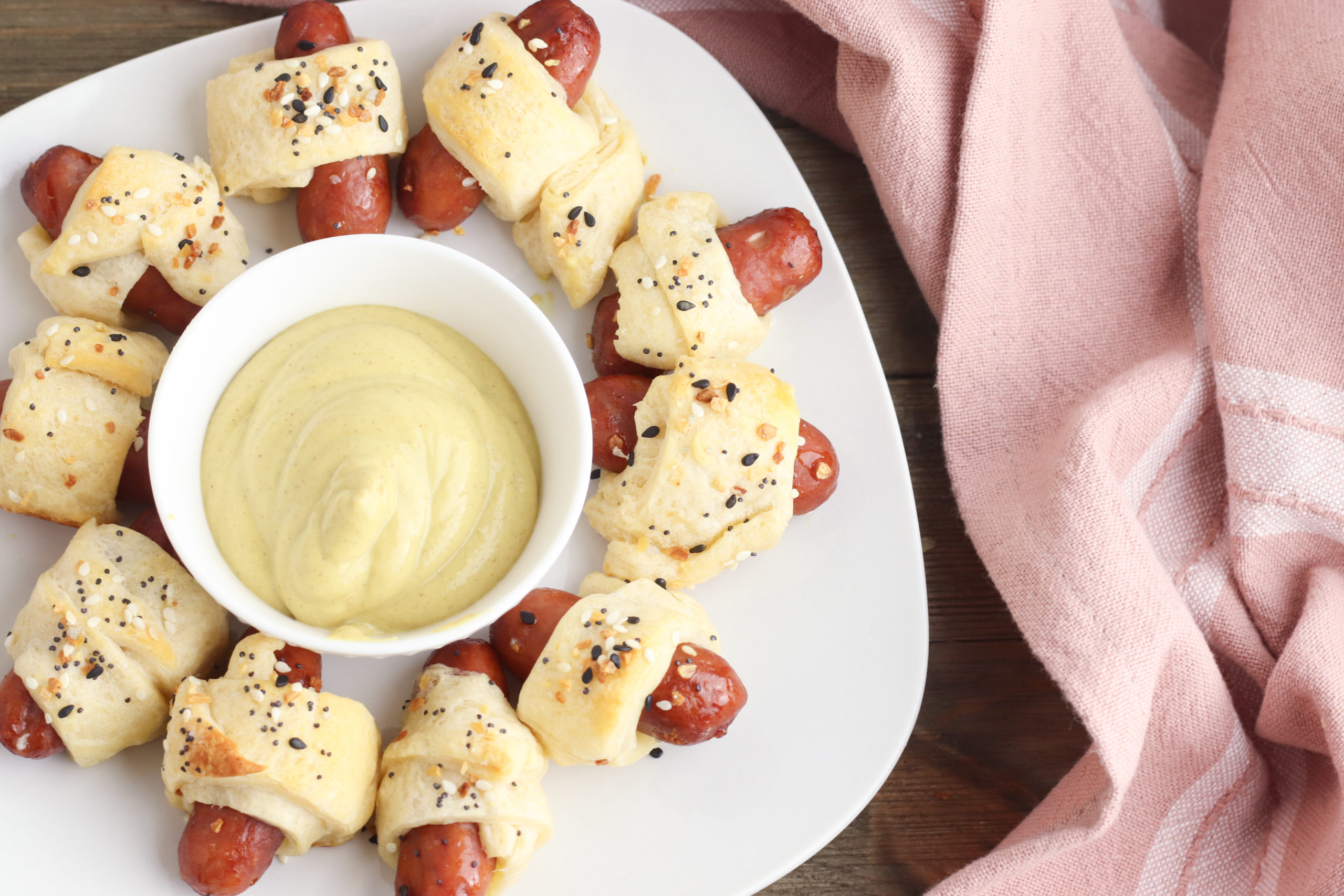 Picture from the top of the pigs in the blanket on a  white tray with a side of dipping sauce and a pink napkin.