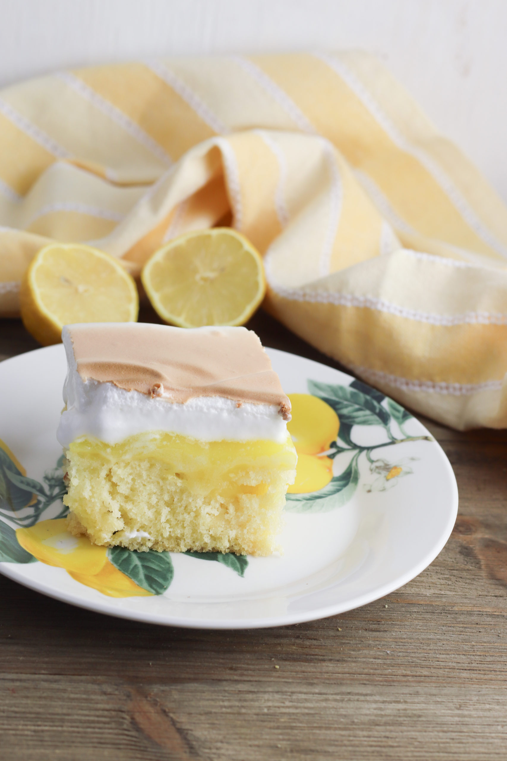 Lemon Meringue poke cake on a white plate with lemons.