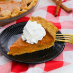 Pie on a plate with whipped cream and a fork on top of a checkered table cloth.
