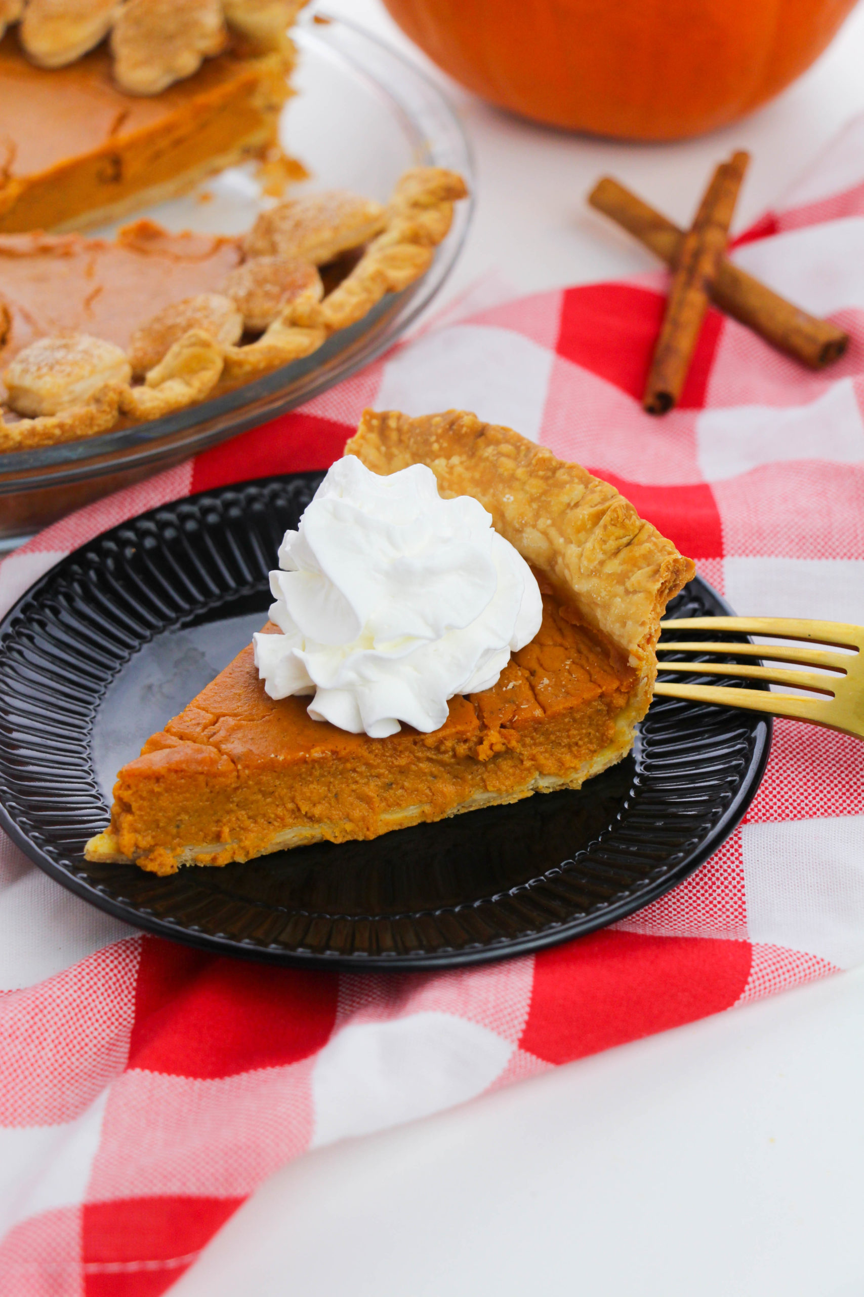 Pie on a plate with whipped cream and a fork on top of a checkered table cloth.
