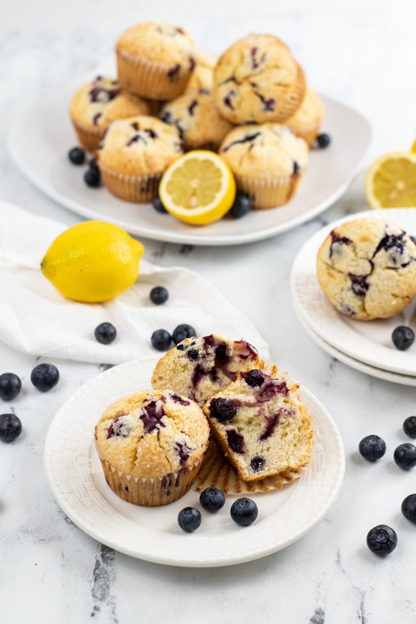 Plate of 3 blueberry lemon muffin with a lemon and a large plate of muffins in the background.